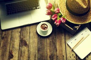 table en bois avec un ordinateur un chapeau des fleurs un carnet avec des crayons à papier et un café